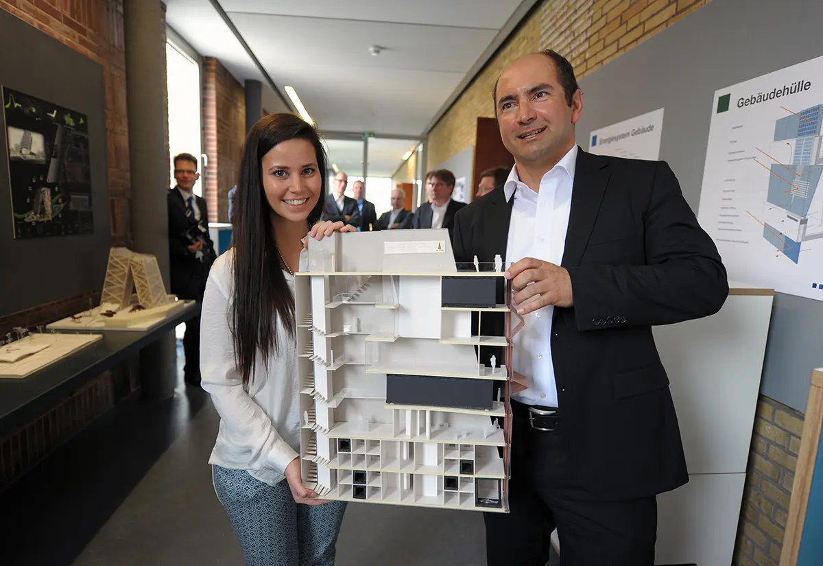 Eine Studentin und ein Professor halten gemeinsam ein Architekturmodell mit Innenansicht in die Kamera. Studiengang Innenarchitektur der h_da, Hochschule Darmstadt.