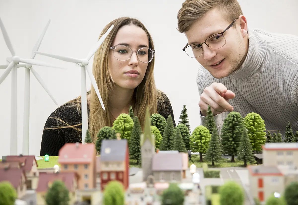 Studierende betrachten ein Modell einer Stadt der Zukunft mit vielen Grünflächen sowie Windrädern. Studiengang Umweltingenieurwesen der h_da, Hochschule Darmstadt.